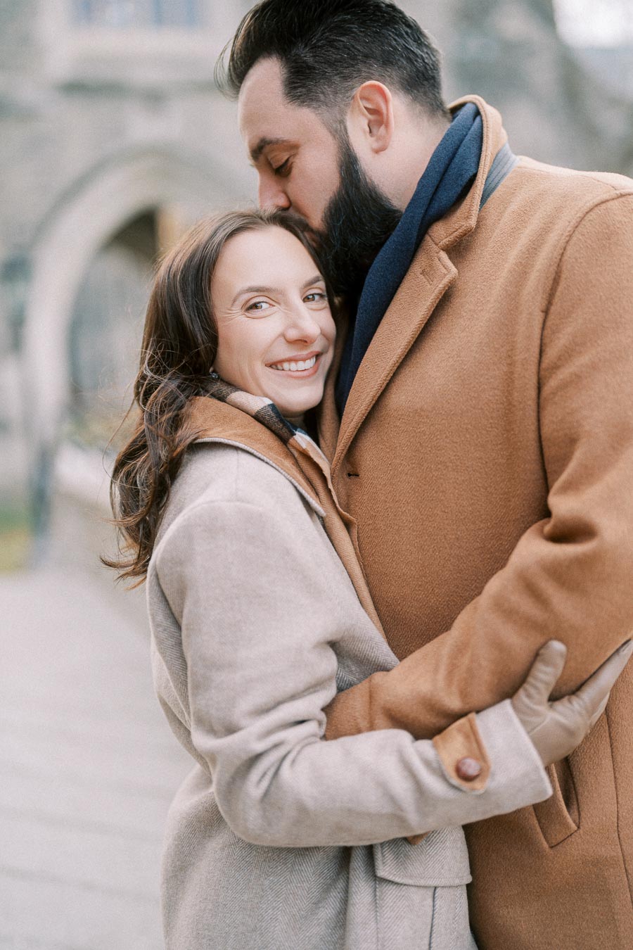 Romantic couple embracing in winter coats, woman smiling while man kisses her forehead, outdoor background.