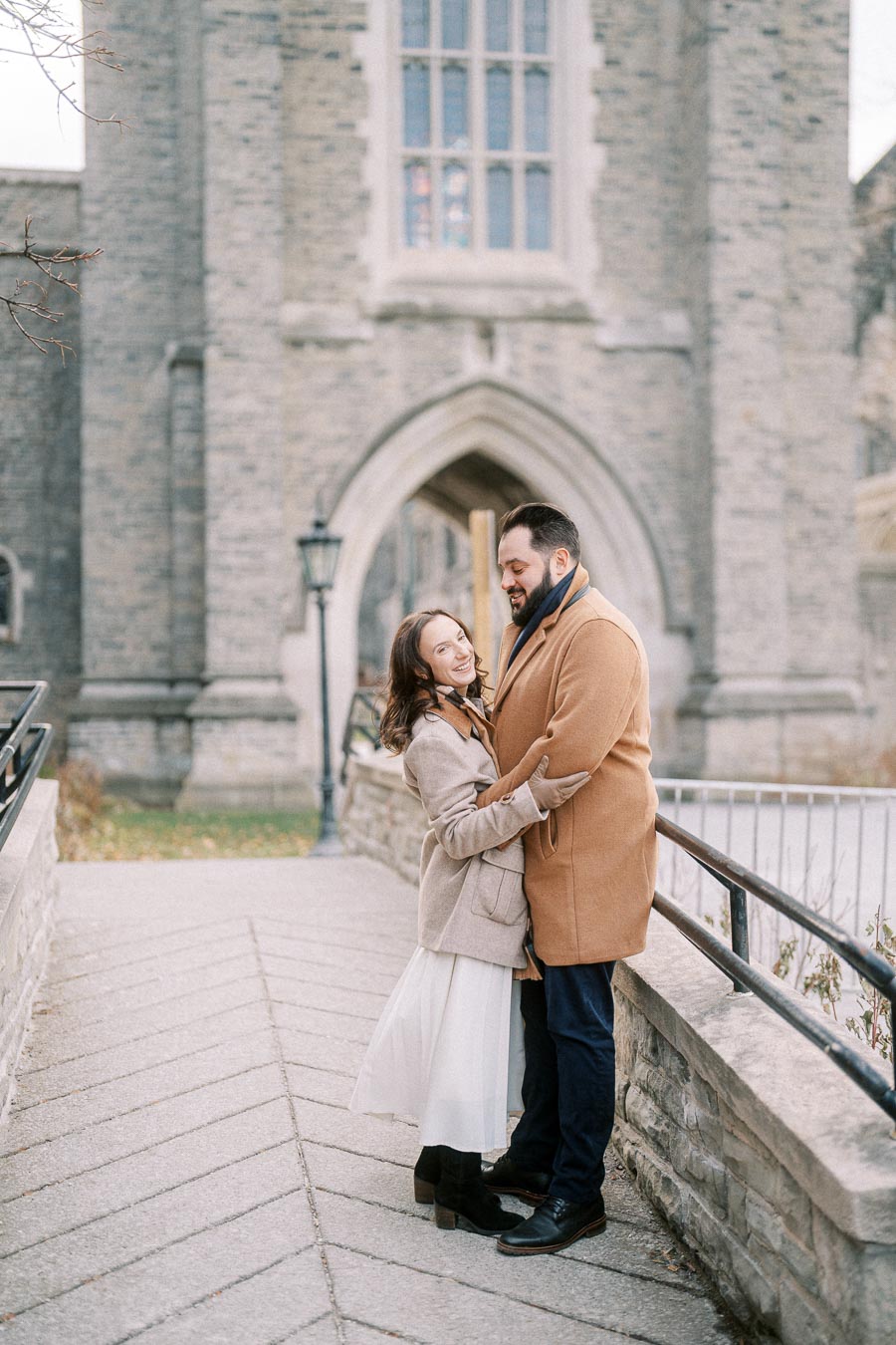 Couple embracing in cozy winter coats outside a historic stone building with an arched window.