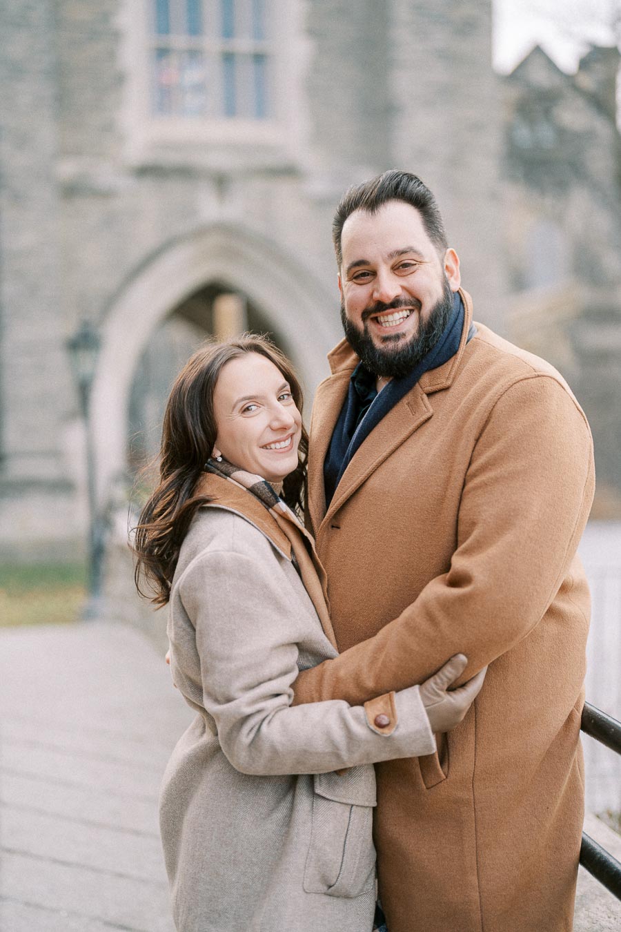 Happy couple embracing outdoors in winter coats, standing in front of a historic stone building with an archway.