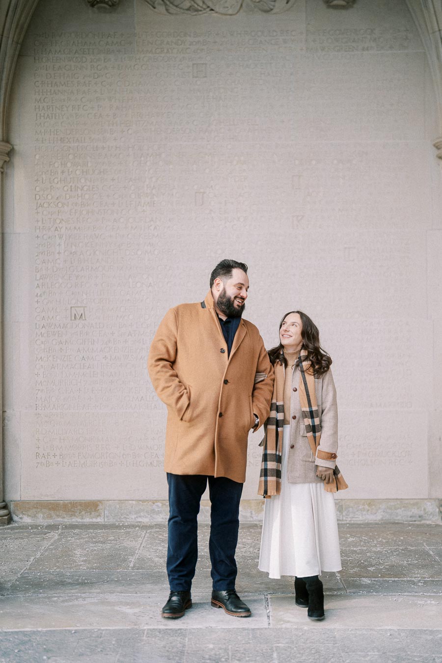 A couple in stylish winter outfits stands smiling in front of a stone wall with engraved text.