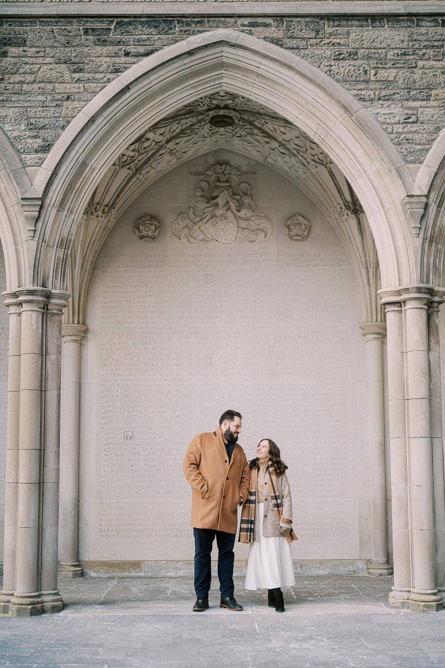 A couple stands smiling under a historic stone archway, wearing stylish winter coats, with textured stonework and inscriptions in the background.