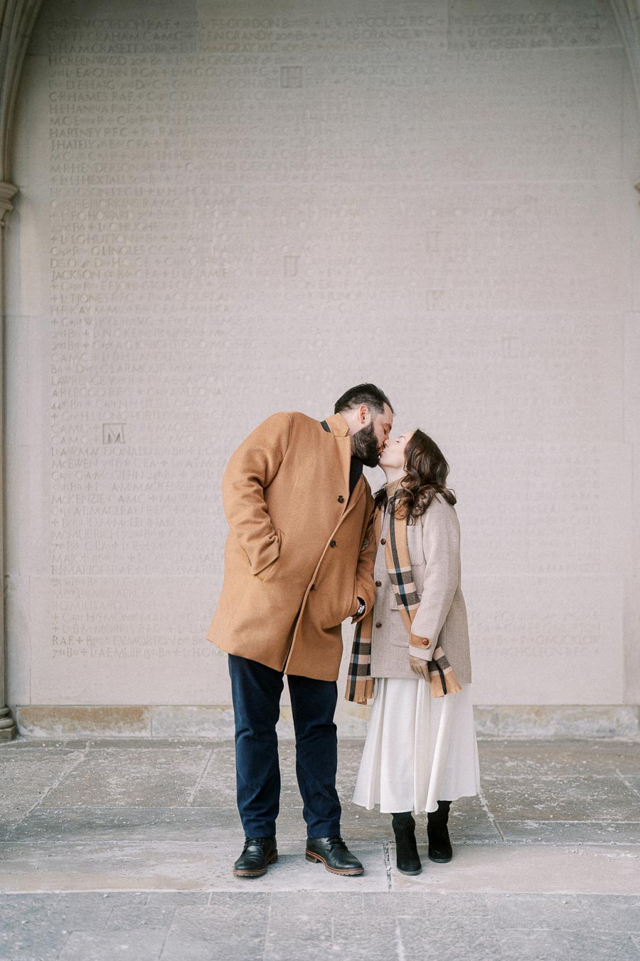 A couple in warm tan coats sharing a kiss, standing in front of a stone wall with engraved text, conveying a romantic moment in an outdoor setting.