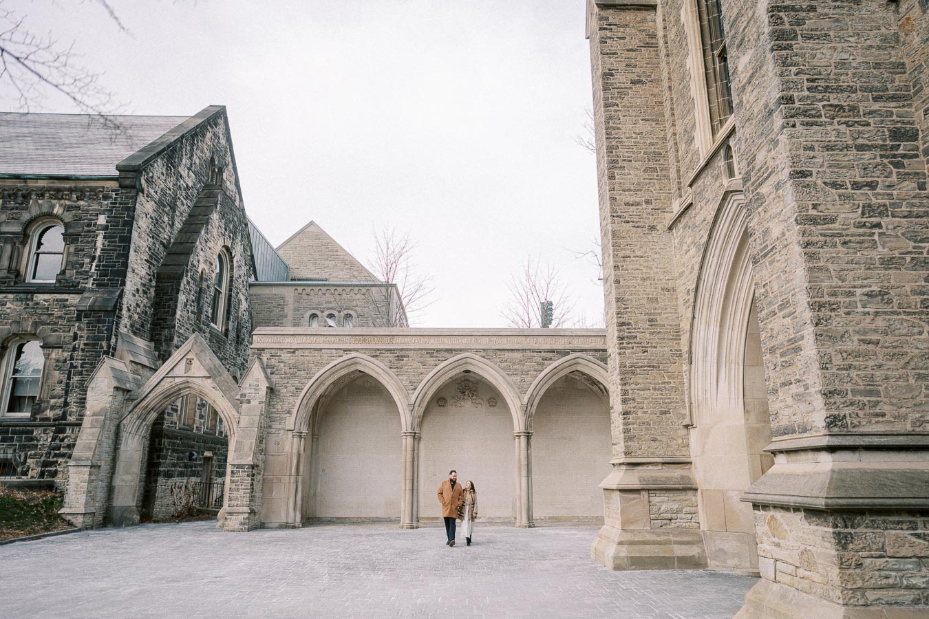 A couple walks through a historic stone courtyard with Gothic arches at a university campus.