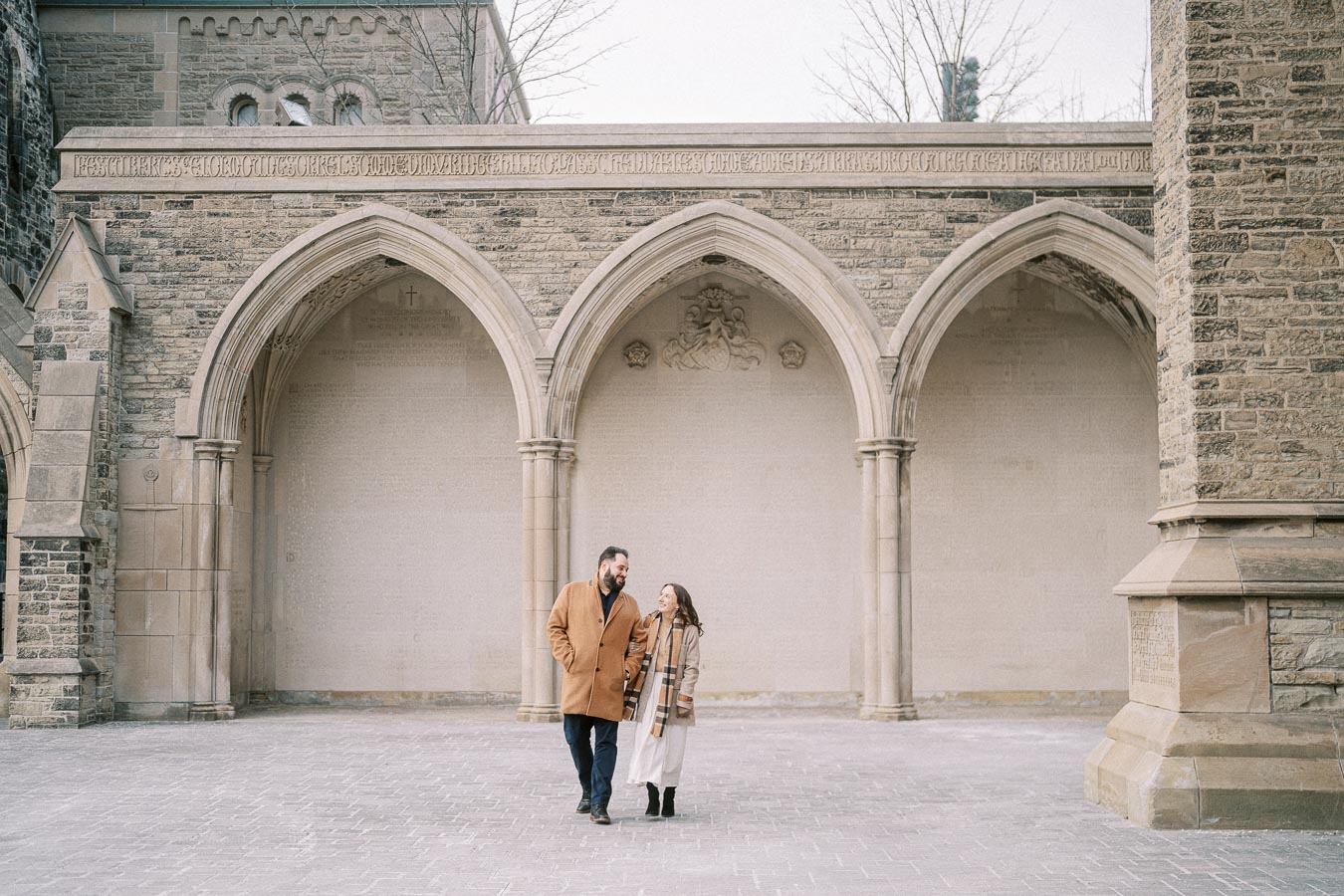 A couple walking and smiling in front of a historic building with stone arches in a peaceful courtyard setting.