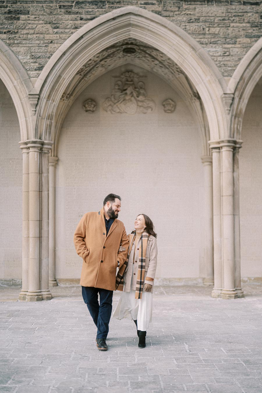 Happy couple walking arm in arm under a gothic archway, wearing stylish winter coats and scarves, smiling at each other in a historic building setting.