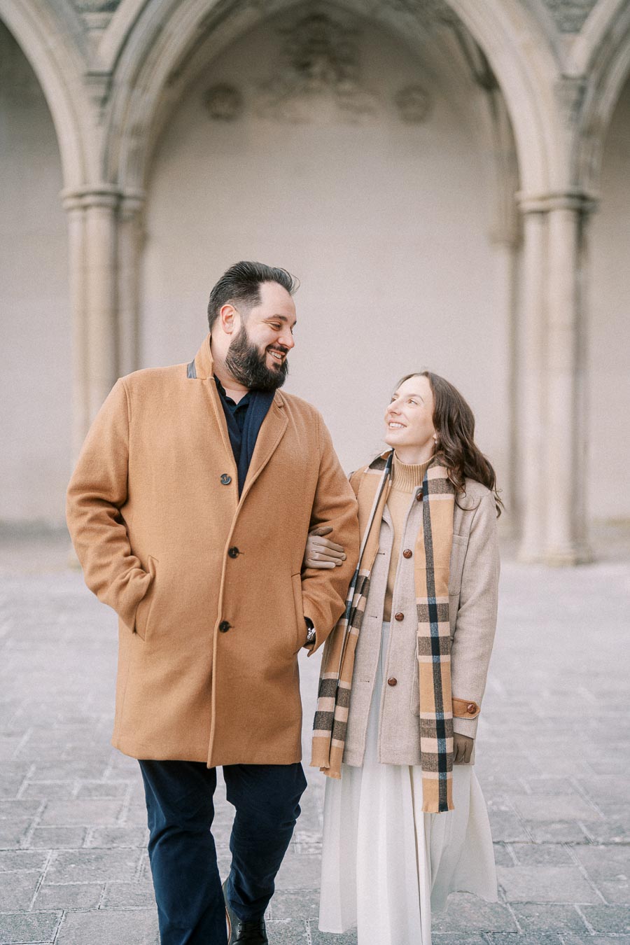 A couple walking together in a stone courtyard, wearing coordinated beige and brown coats with matching scarves, smiling affectionately at each other.