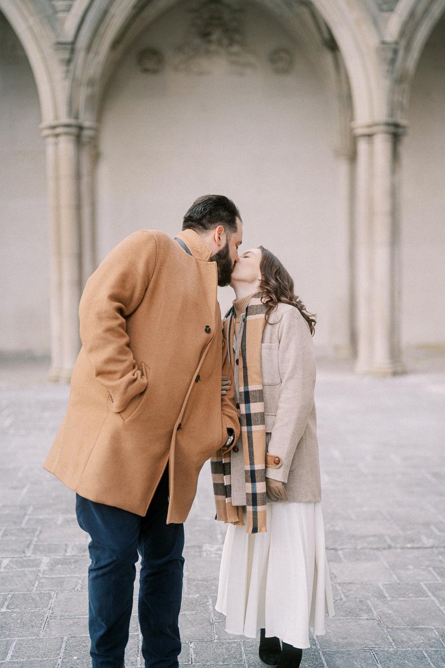 Couple sharing a romantic kiss in stylish winter coats and scarf, standing under an ornate stone archway.