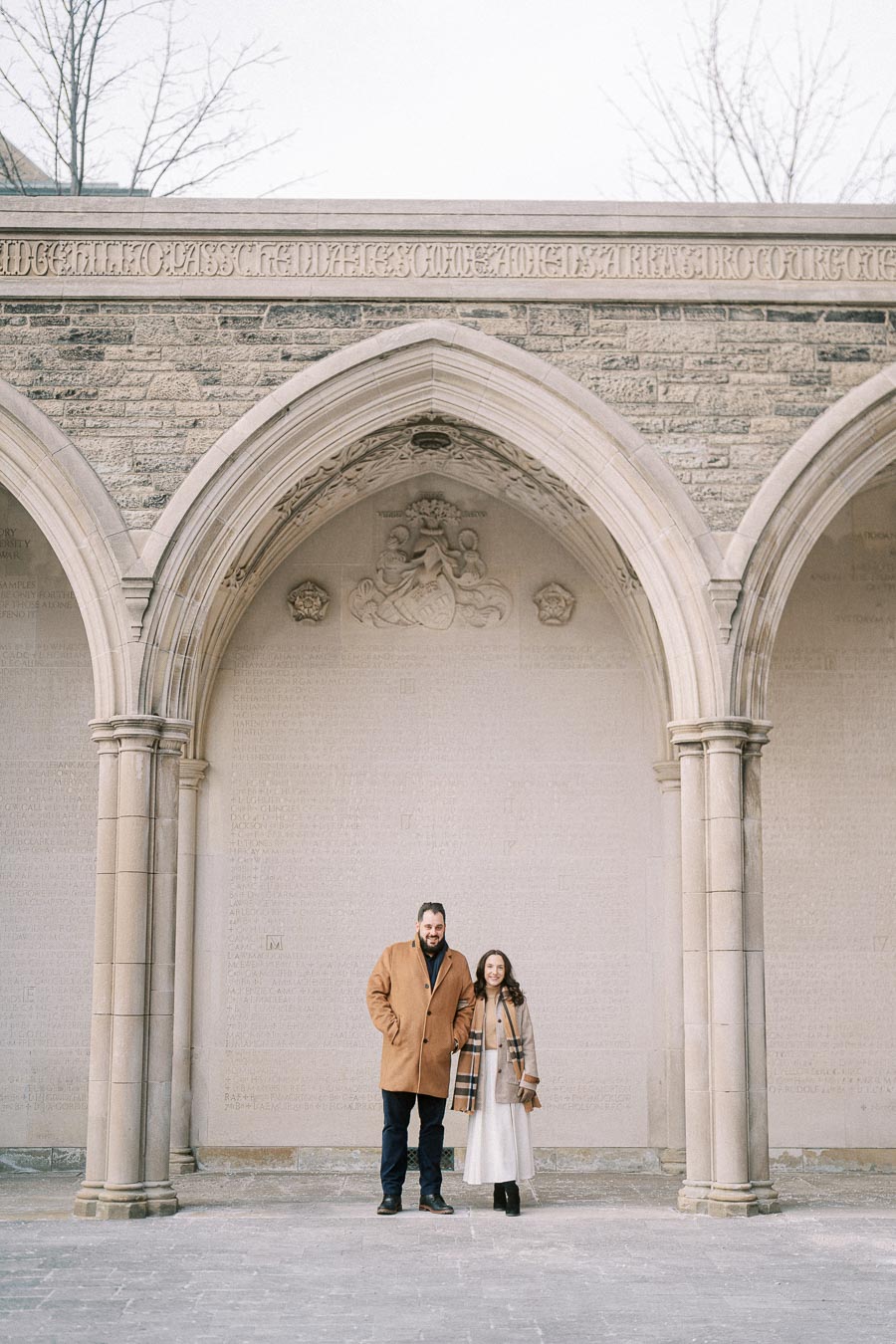 Couple standing under historical stone archway, dressed in winter coats, with engraved wall and bare trees in the background.