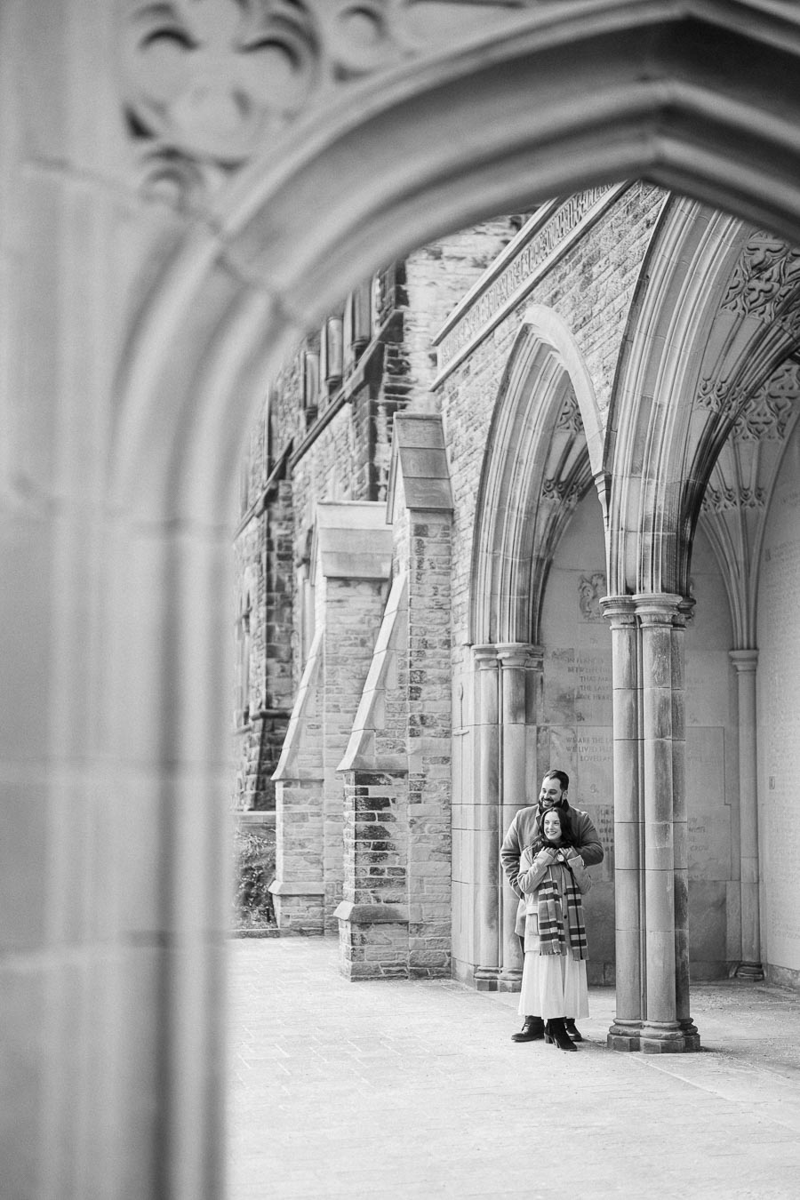 A couple stands embracing under historic stone arches, showcasing intricate Gothic architectural details in black and white photography.
