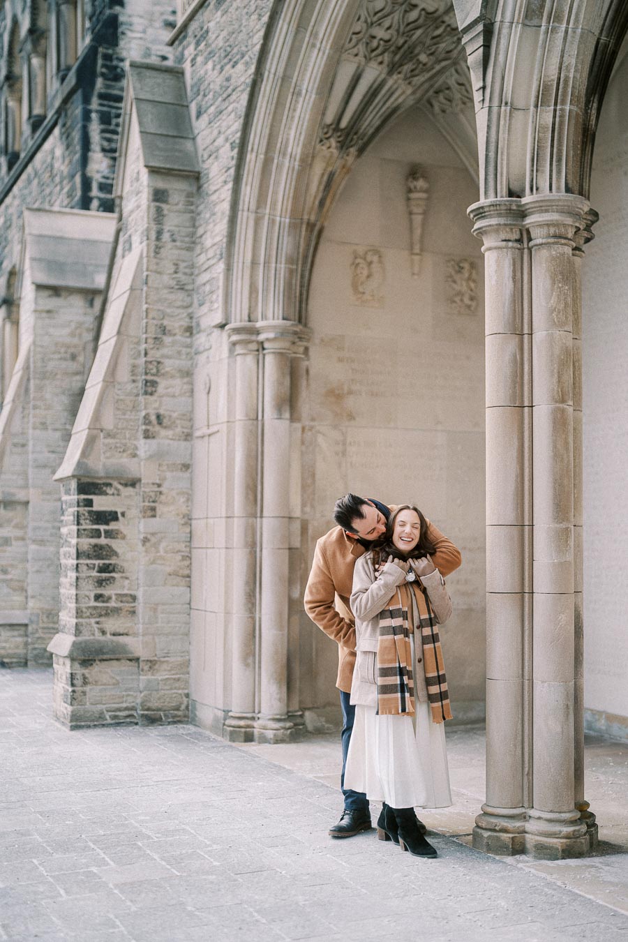 Happy couple embracing near historic stone architecture, wearing winter coats and scarves, showcasing a cozy romantic moment.