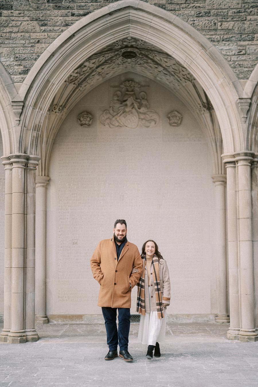 A couple stands happily under a historic stone archway, both wearing warm coats and smiling, with intricate carvings visible in the background.