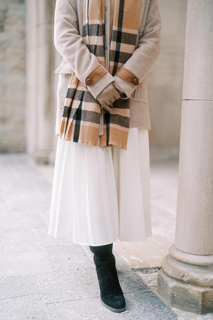 Person wearing a beige coat and plaid scarf with black boots and a white skirt, standing beside a stone pillar.