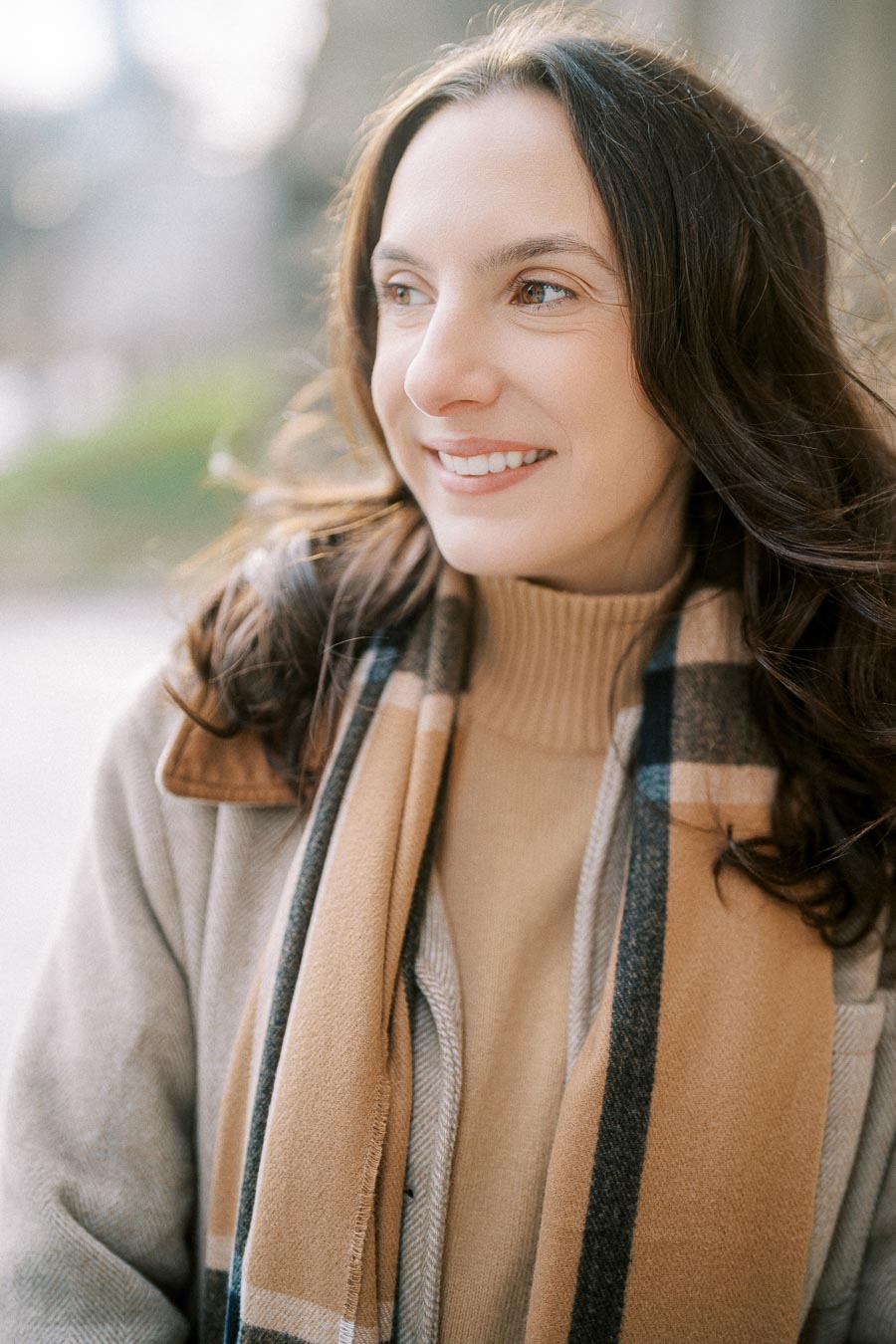 Portrait of a woman smiling in a winter setting, wearing a beige scarf and coat, with natural light highlighting her face, showcasing a serene outdoor atmosphere.