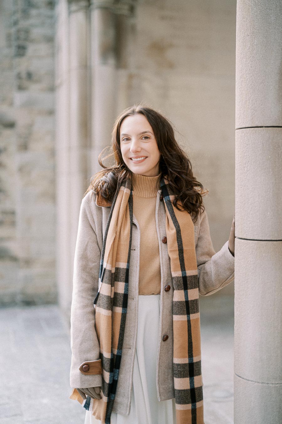 A woman in a stylish tan coat and plaid scarf smiles while standing outside against a stone building background.