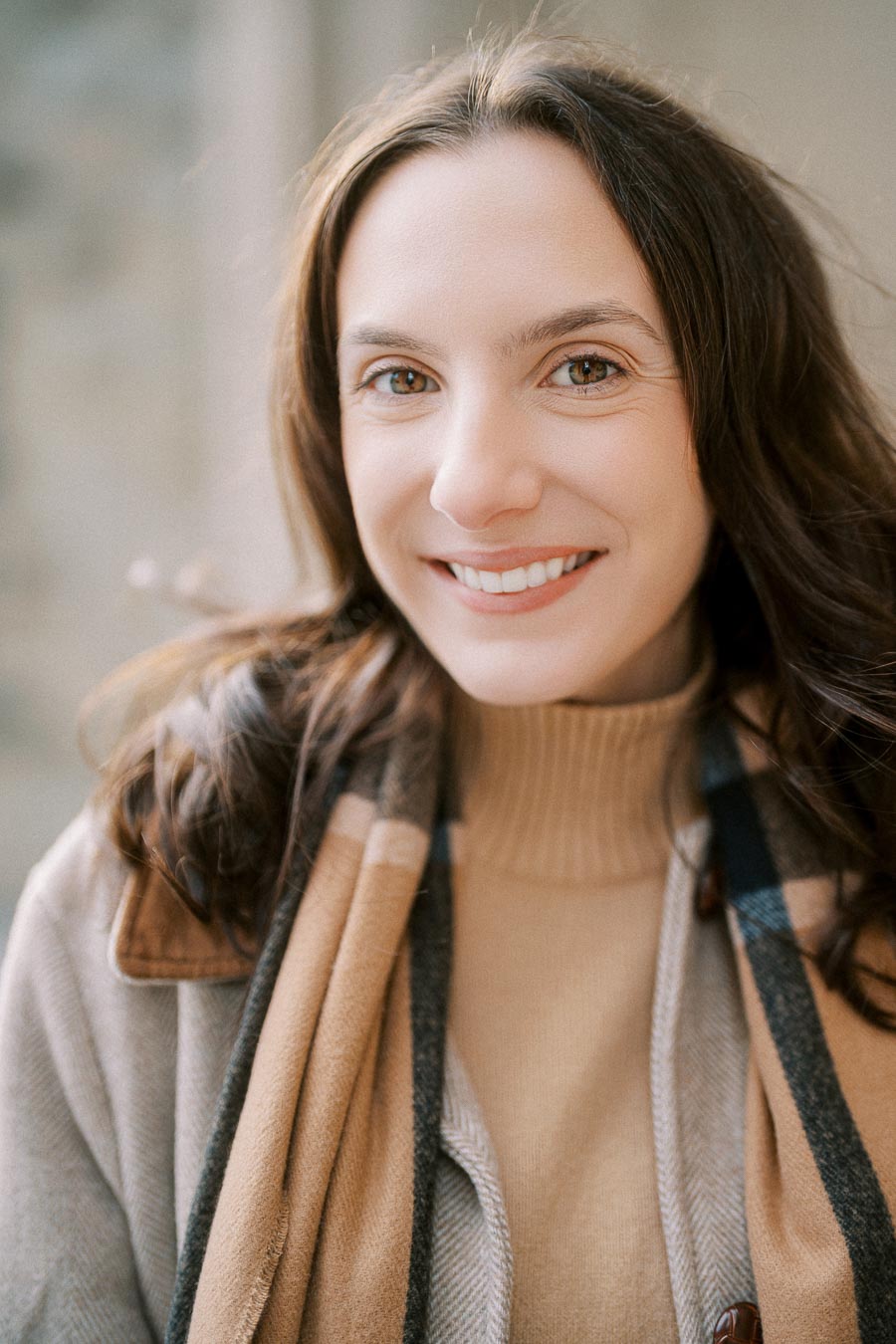 Smiling woman with long brown hair wearing a cozy scarf and beige sweater, standing outdoors
