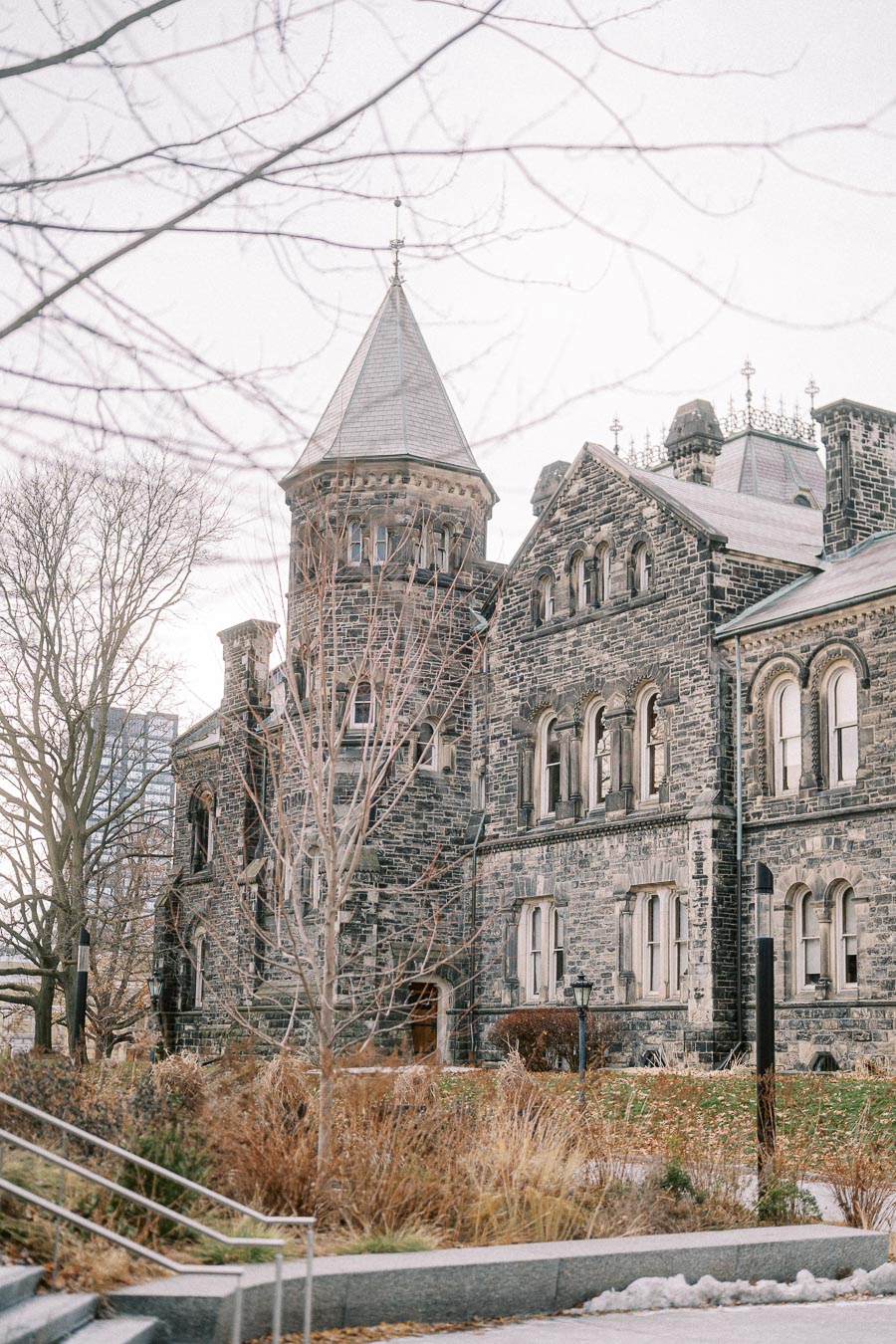 Historic Gothic-style stone building with a spire, surrounded by bare winter trees and a walkway in the foreground.