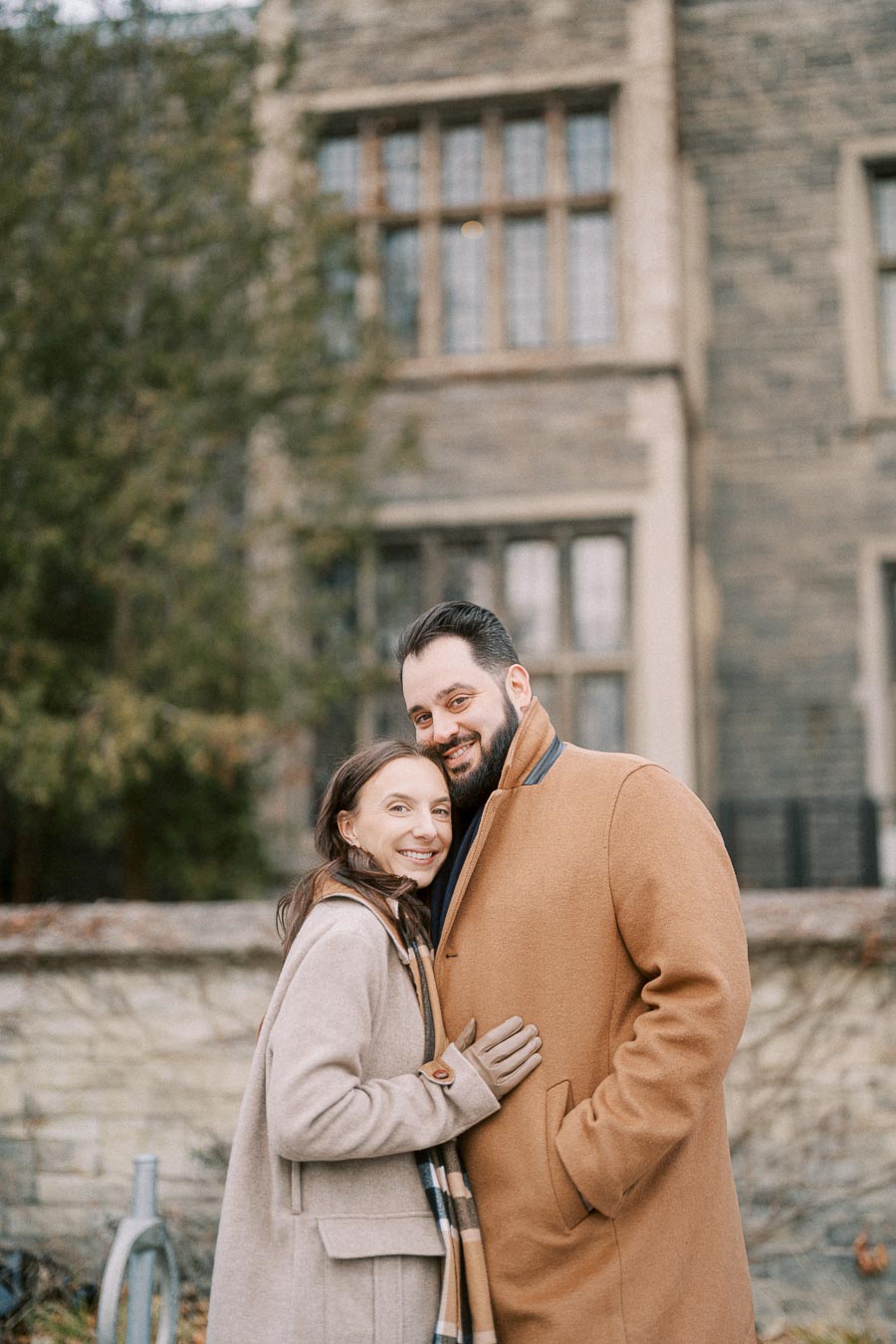 Smiling couple embracing outdoors, wearing stylish winter coats with a historical building backdrop