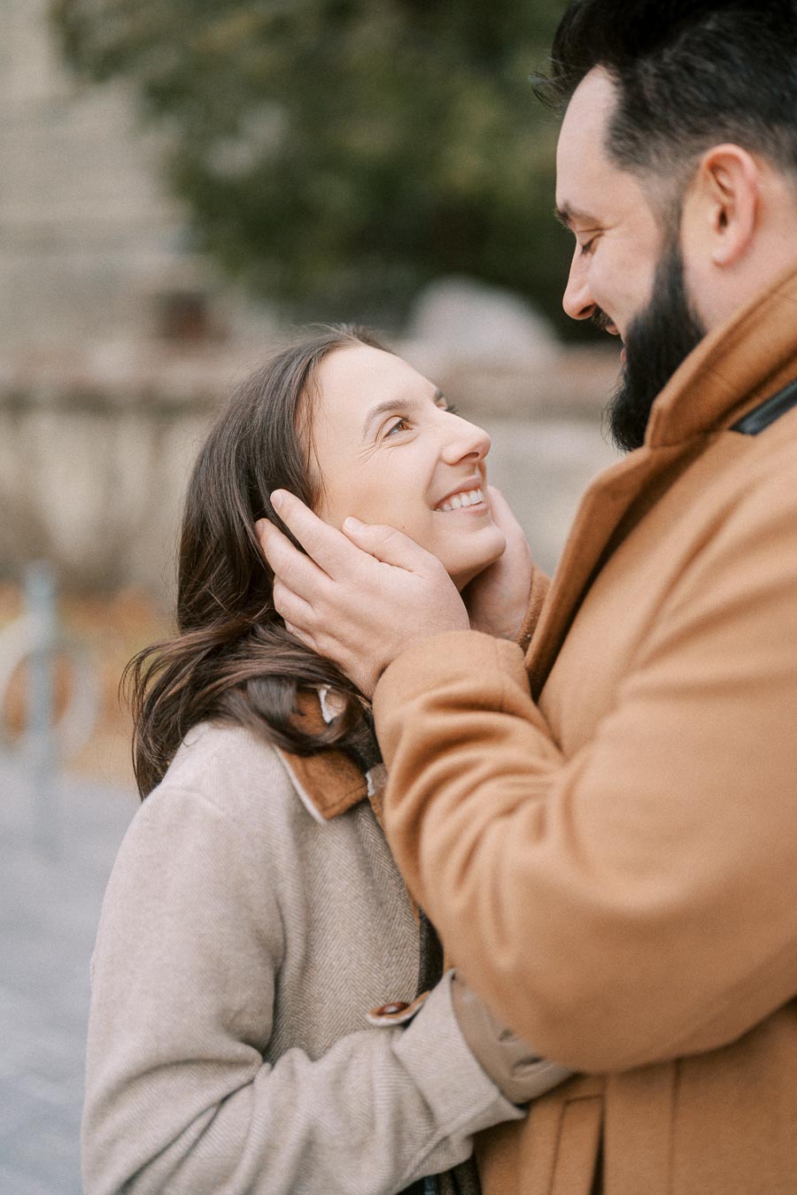 A happy couple shares a romantic moment outdoors, with the man gently holding the woman's face and both smiling warmly at each other. They're dressed in cozy, neutral-toned winter coats with a blurred background of trees and a sidewalk.