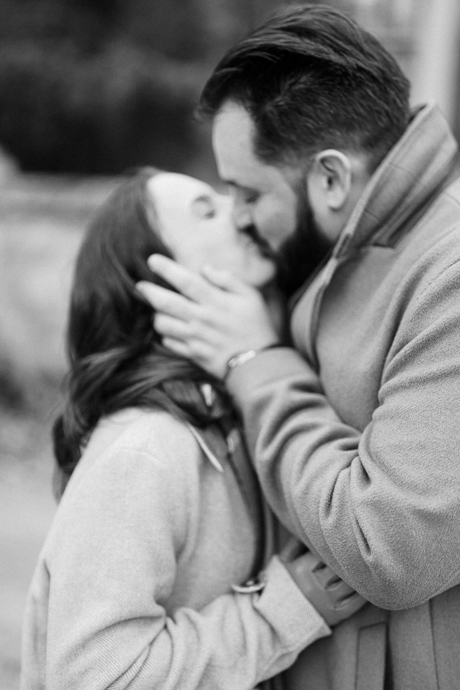 A black and white photograph of a couple kissing outdoors, both wearing winter coats, capturing a tender and intimate moment.