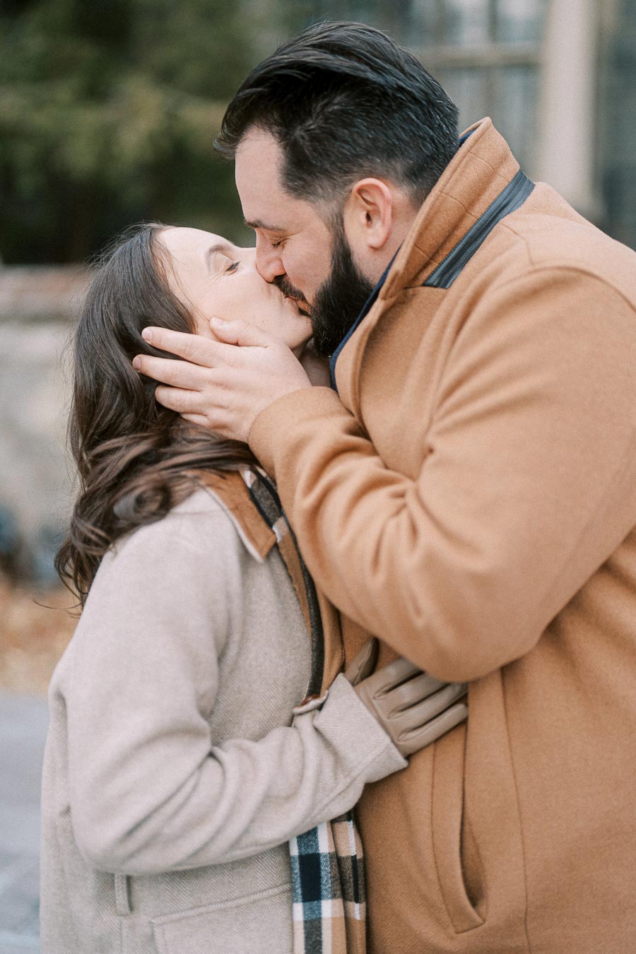 A couple sharing a romantic kiss outdoors, both wearing warm beige coats, capturing a heartfelt winter moment.