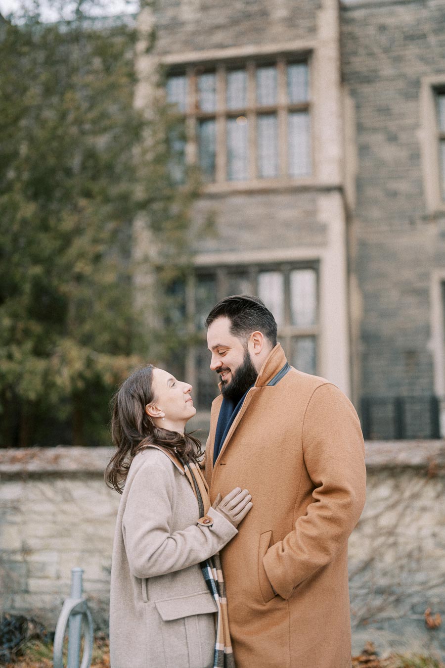 A couple in winter coats smiling at each other outside a historic stone building, surrounded by greenery.