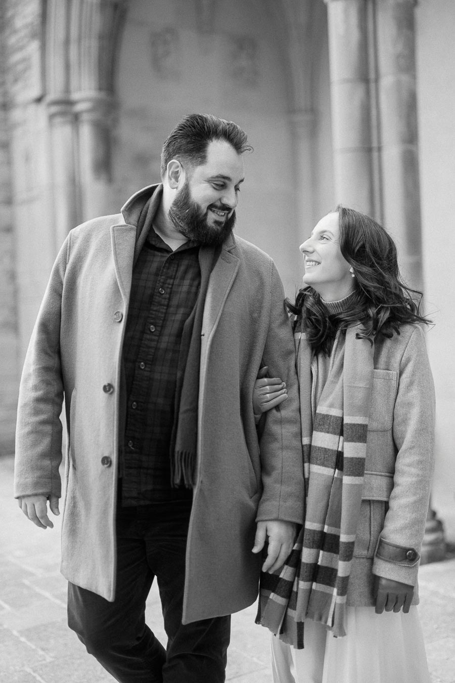 A black and white photo of a smiling couple walking arm-in-arm, dressed in winter coats and scarves, with an elegant stone archway in the background.