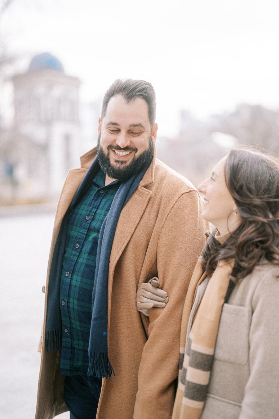 A smiling couple in warm winter coats and scarves walking arm in arm, enjoying a light-hearted moment outdoors in a city setting.