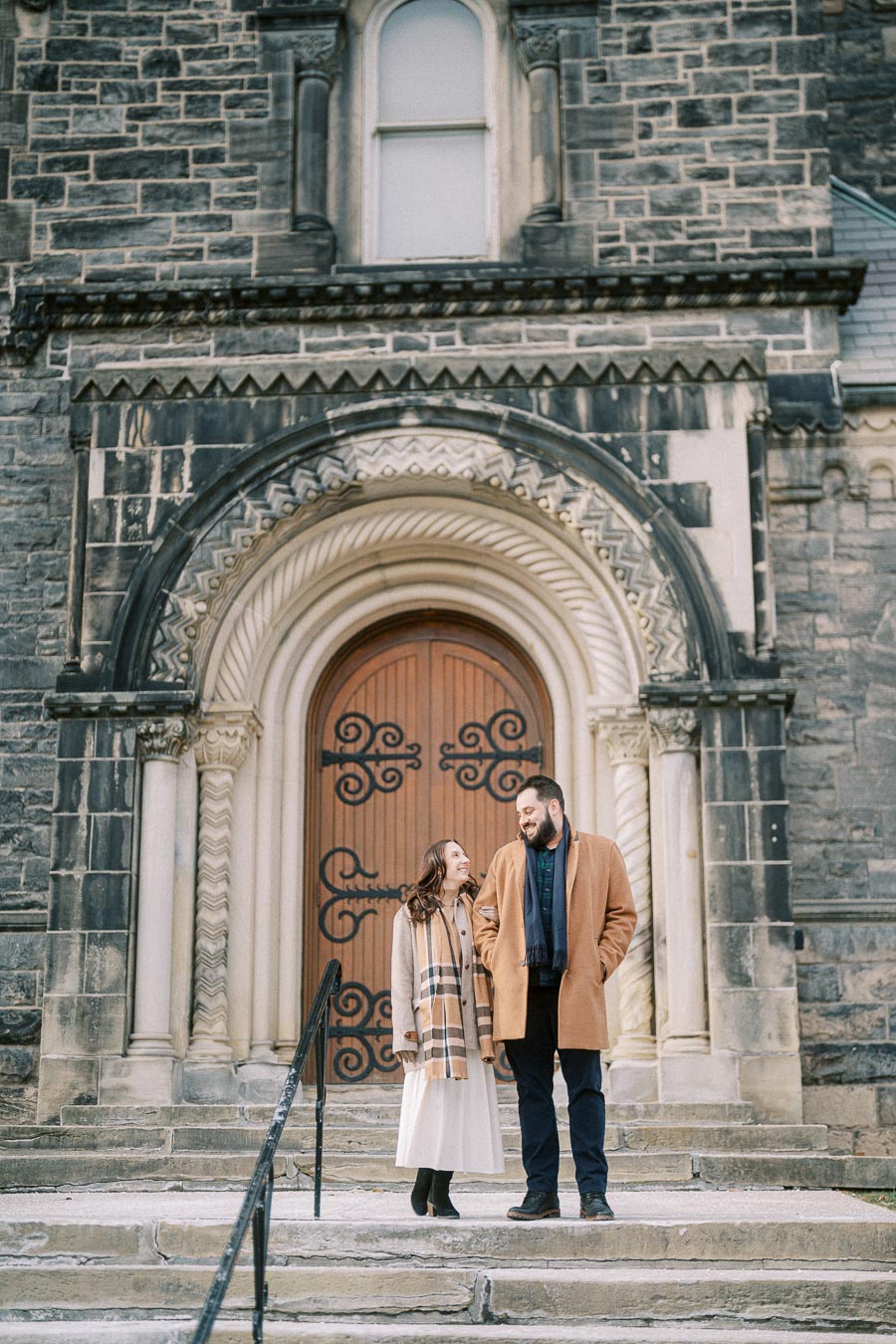 A couple in winter coats smiling at each other while standing on the stone steps of a historic building with ornate wooden doors.