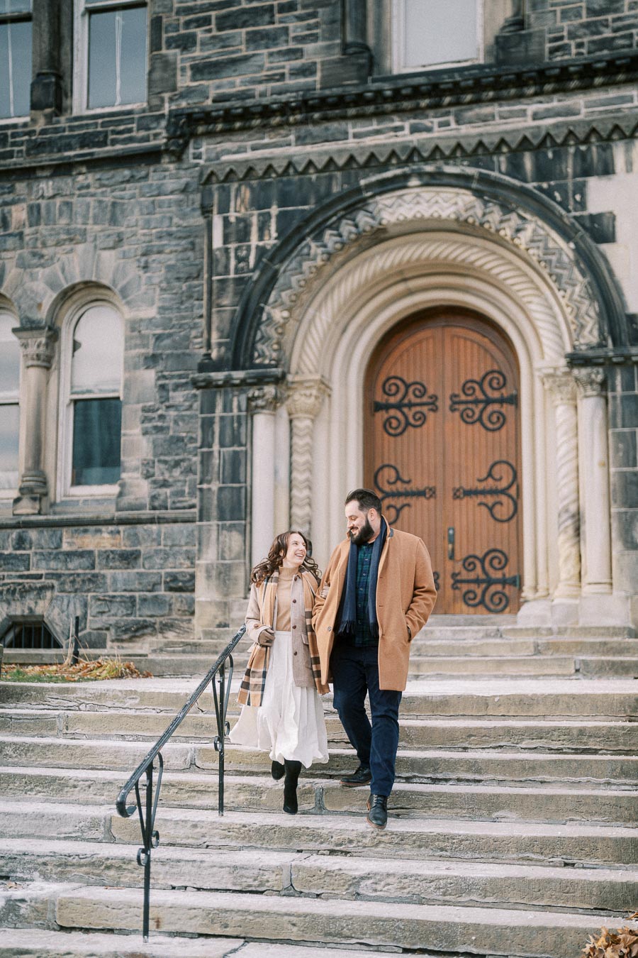 Couple in stylish winter coats walking down stone steps of historic building with ornate wooden door.