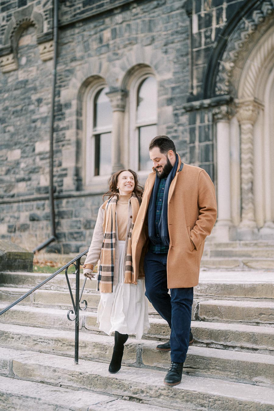 A couple in stylish winter attire walking down stone steps in front of a historic stone building, with the woman wearing a beige coat and scarf and the man in a brown overcoat.