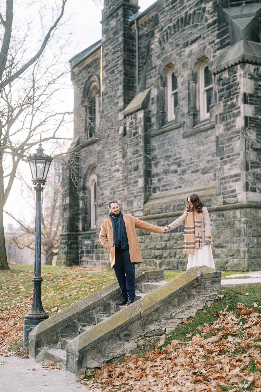 A couple holding hands and walking down stone steps in front of a historic stone building during autumn.