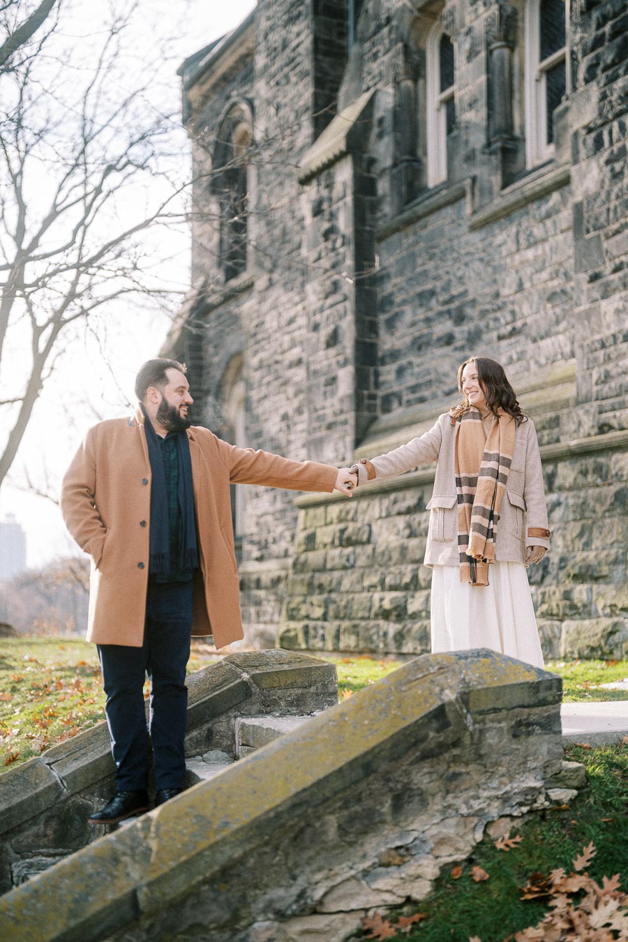 Couple holding hands near historic stone building, both wearing beige coats, enjoying a sunny autumn day.