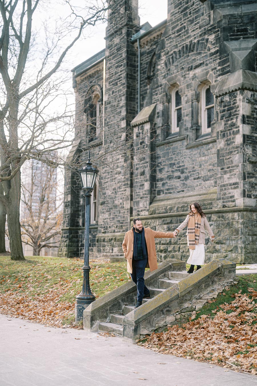 A couple walking hand in hand down stone steps in front of a historic stone building during autumn, surrounded by fallen leaves and bare trees.
