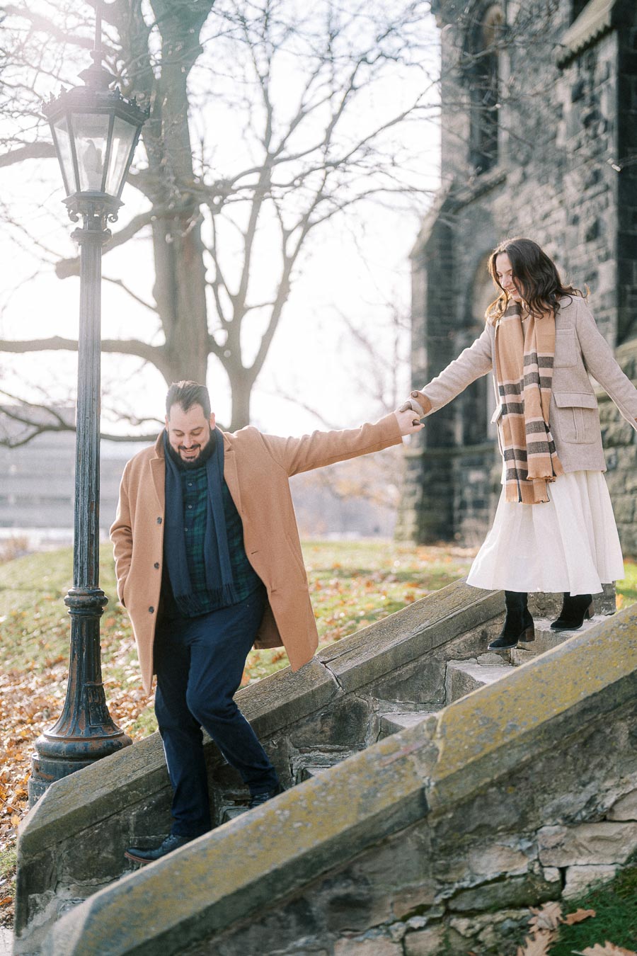 A couple in stylish winter coats walking hand in hand down stone steps next to an old building, with autumn leaves scattered around.