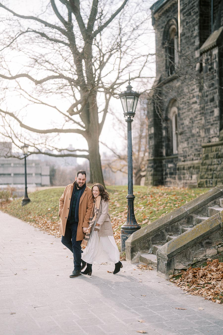 Couple walking and laughing on a sidewalk beside a historic stone building and lamp post, with bare trees and fallen leaves in the background.