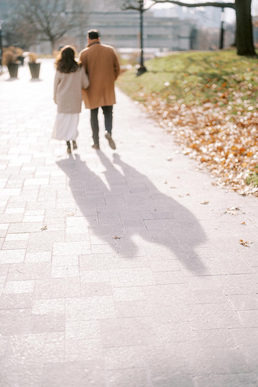 A couple walking down a sunlit path in autumn, wearing warm coats, with leaves scattered along the sidewalk.