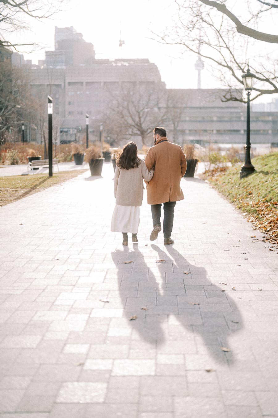 A couple walks arm in arm down a sunlit city park path on a clear autumn day, surrounded by bare trees and urban architecture in the background.