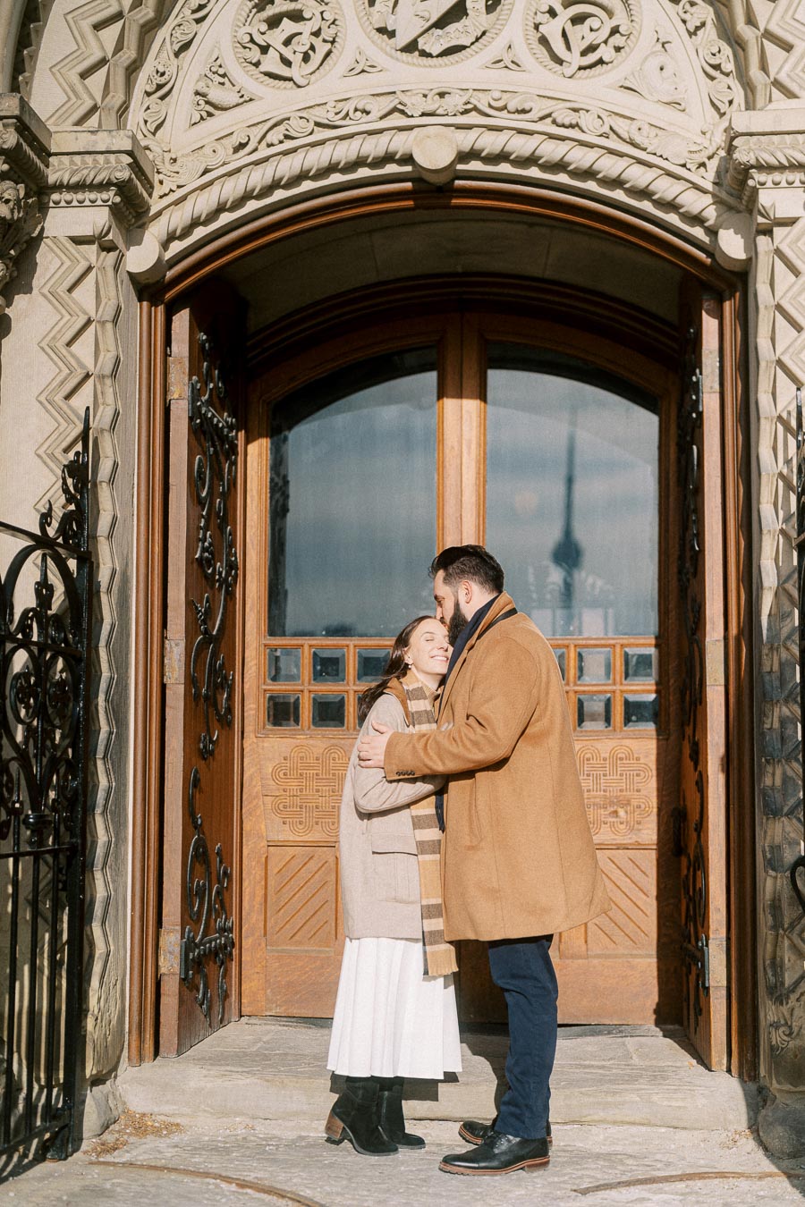 Couple embracing in front of ornate wooden door, wearing stylish winter coats, with intricate stone architecture and wrought iron details visible.