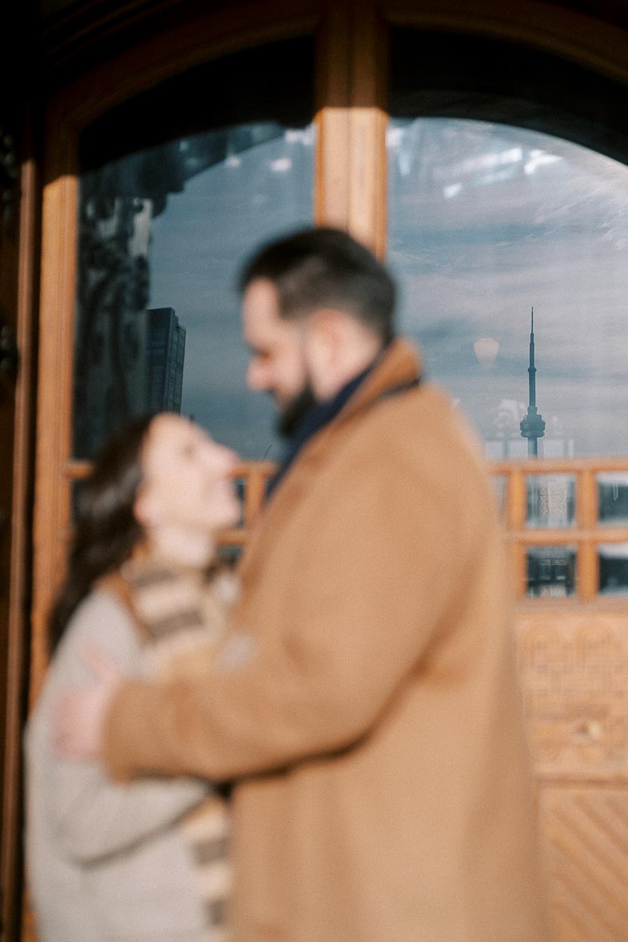 Blurry couple embracing with view of iconic CN Tower reflected in a window, capturing a romantic moment in Toronto.