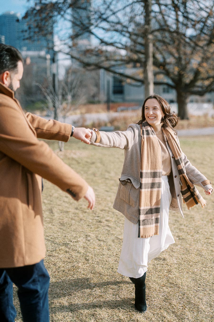 A couple in warm winter clothing enjoying a playful outdoor moment holding hands in a park, with city buildings and bare trees in the background.