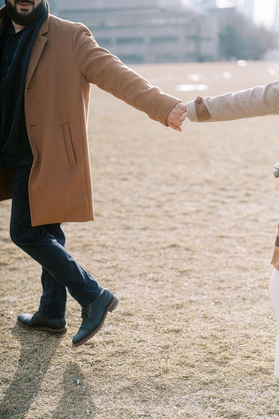A person in a brown coat holding hands with another person in a light coat, walking on a grass field with a blurred building in the background.