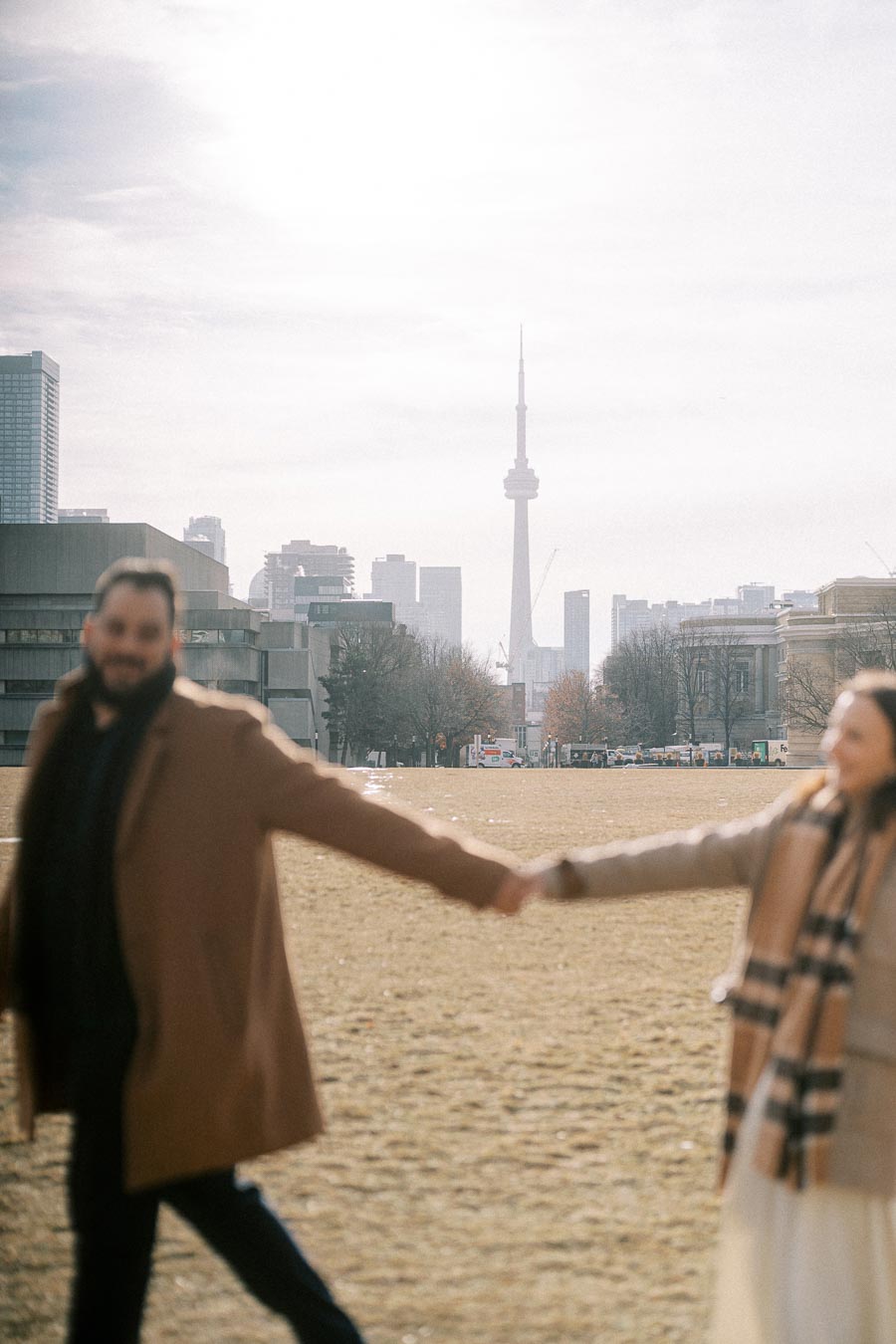 A couple holds hands in a blurred foreground with the iconic CN Tower and Toronto skyline visible in the background on a sunny day.