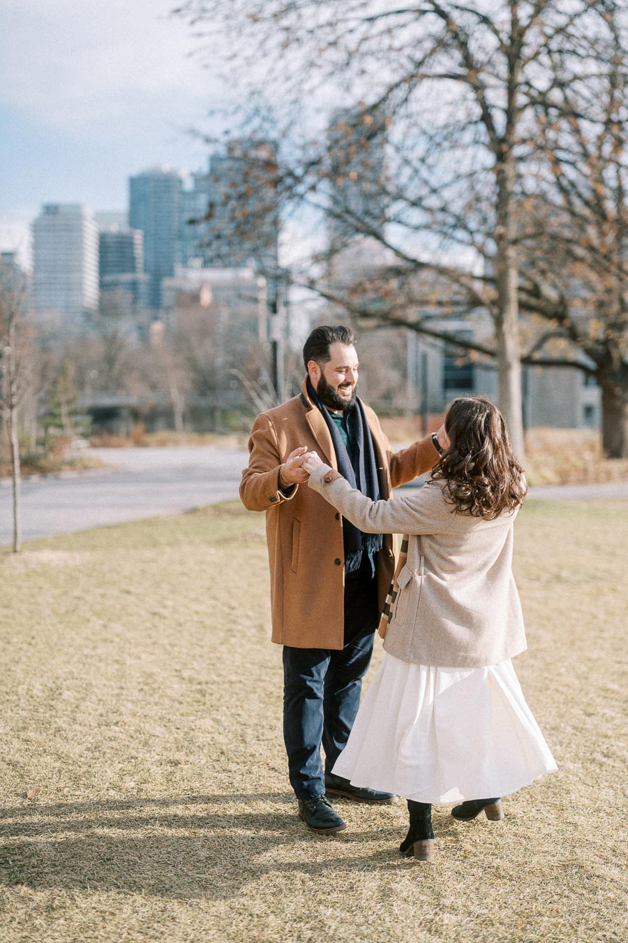 A couple dancing in a park on a sunny day with city skyline in the background, wearing winter coats and smiling warmly at each other.
