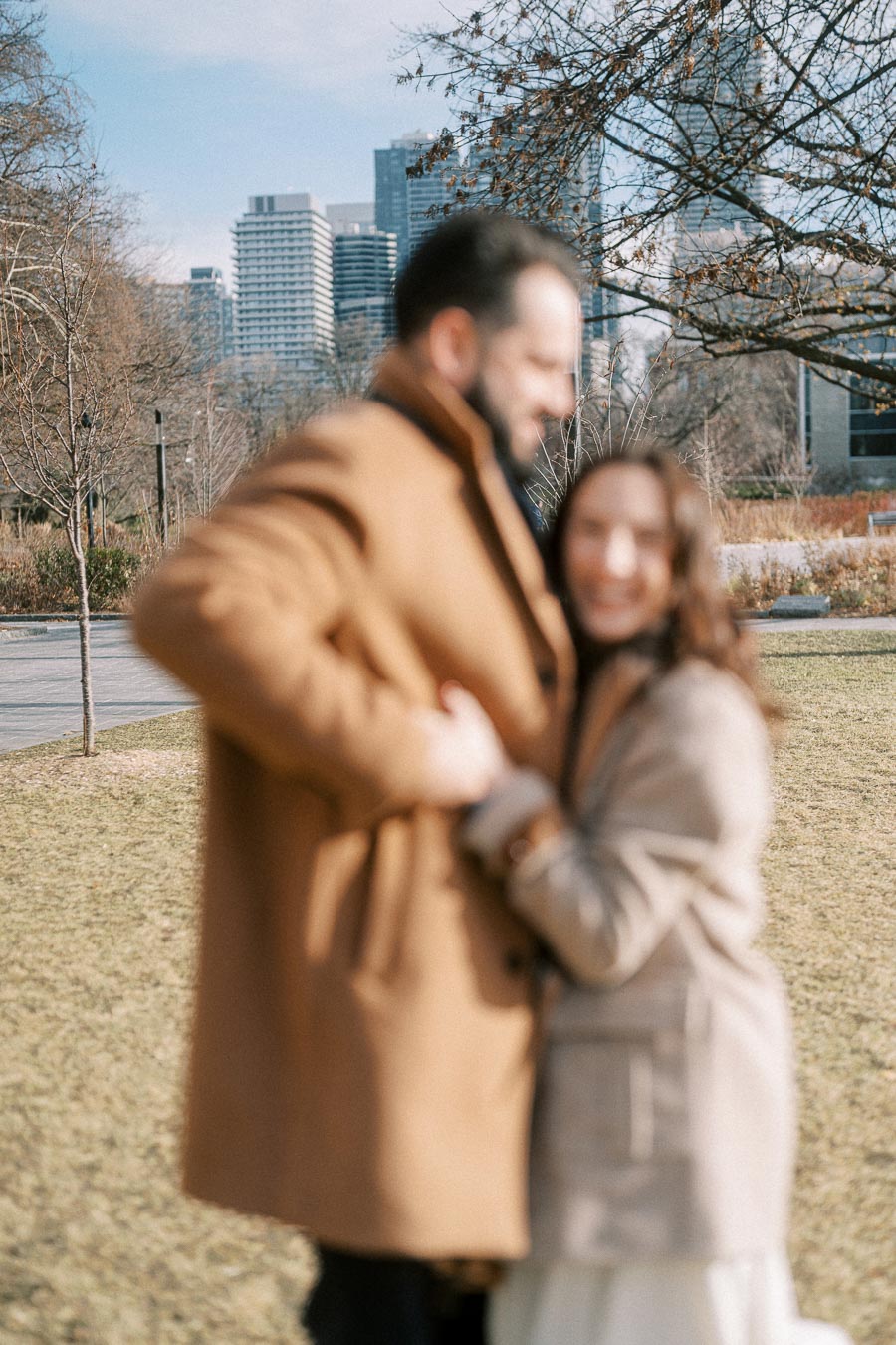 Blurry photo of a couple embracing outdoors in winter coats, with a cityscape background featuring high-rise buildings and bare trees.