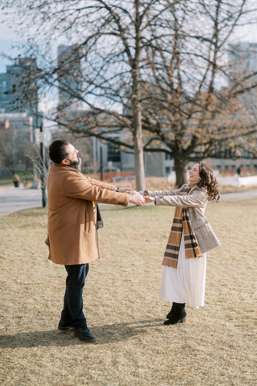 A couple joyfully holding hands and spinning in a park on a sunny day, with bare trees and an urban skyline in the background, both wearing stylish winter coats and scarves.