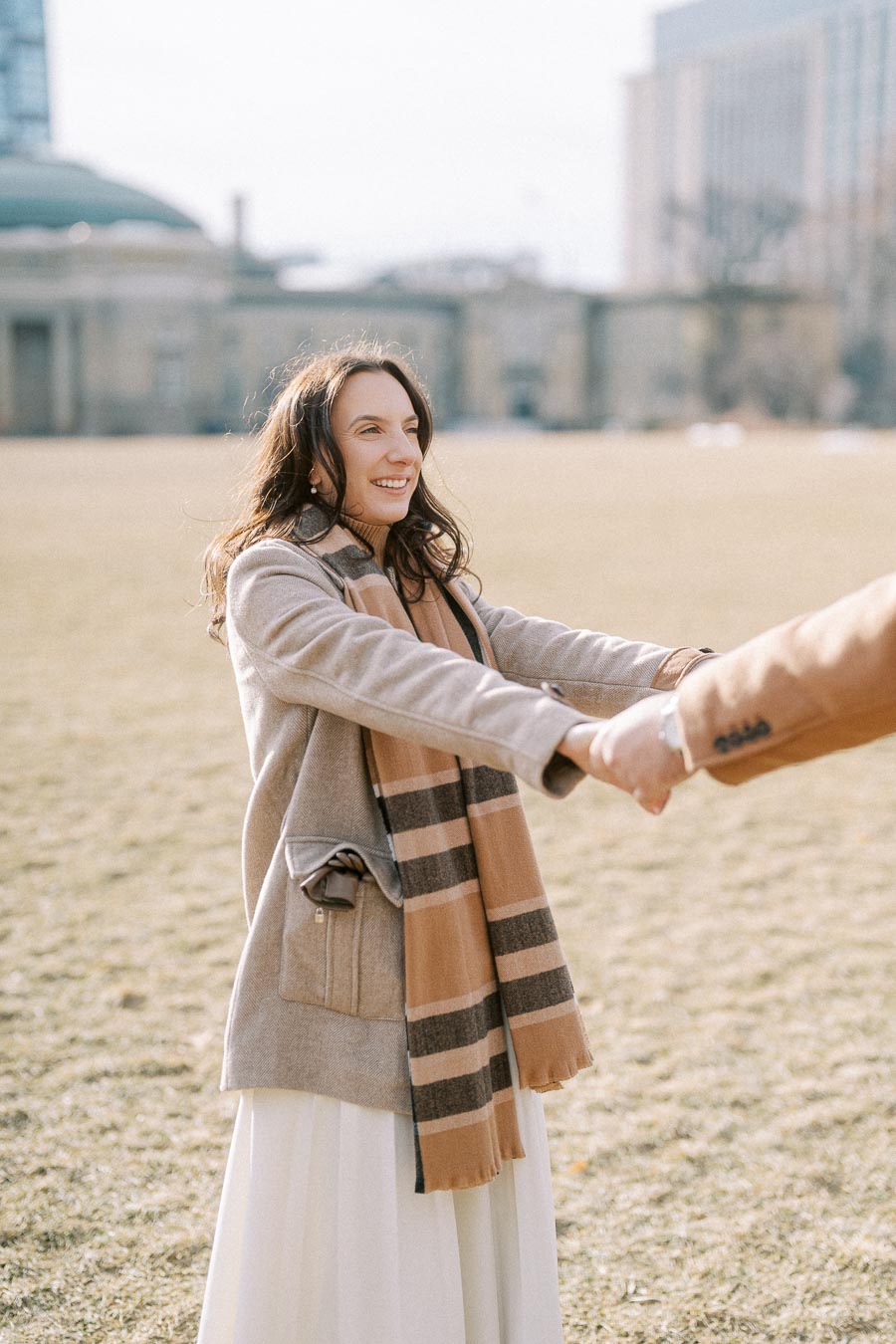 Smiling woman in a beige coat and scarf holding hands in a park during a sunny day.
