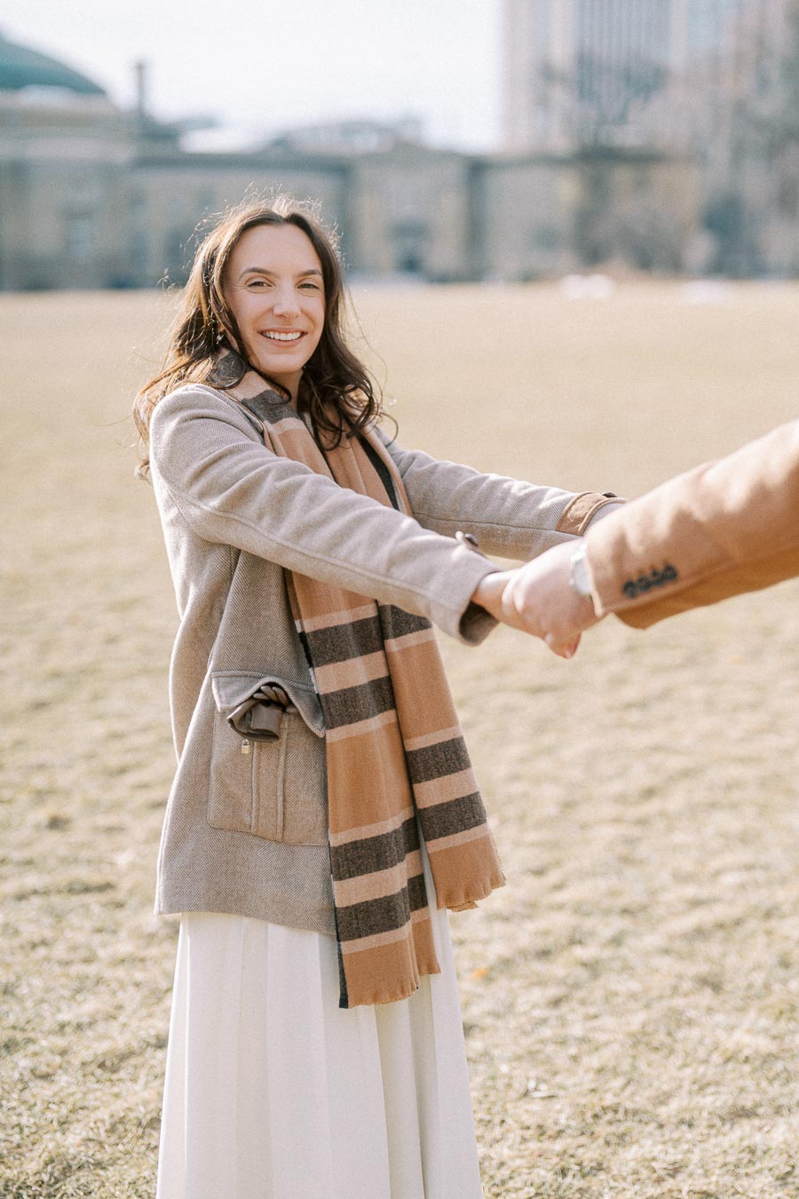 Smiling woman in a winter coat and scarf holding hands outdoors in a sunny park setting.