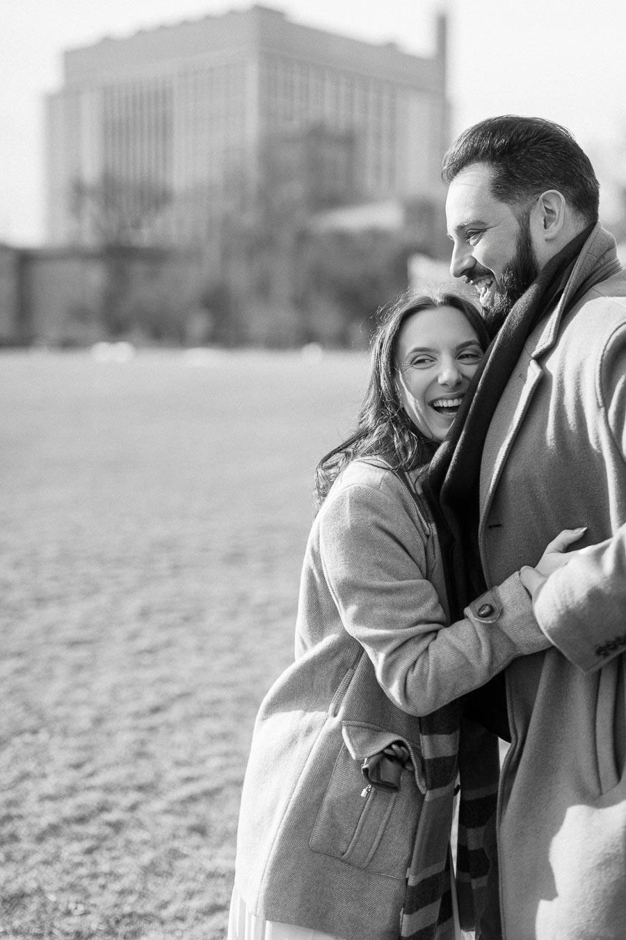 A happy couple smiling and embracing in an outdoor park setting, dressed warmly in overcoats and scarves, with a large building in the blurred background.