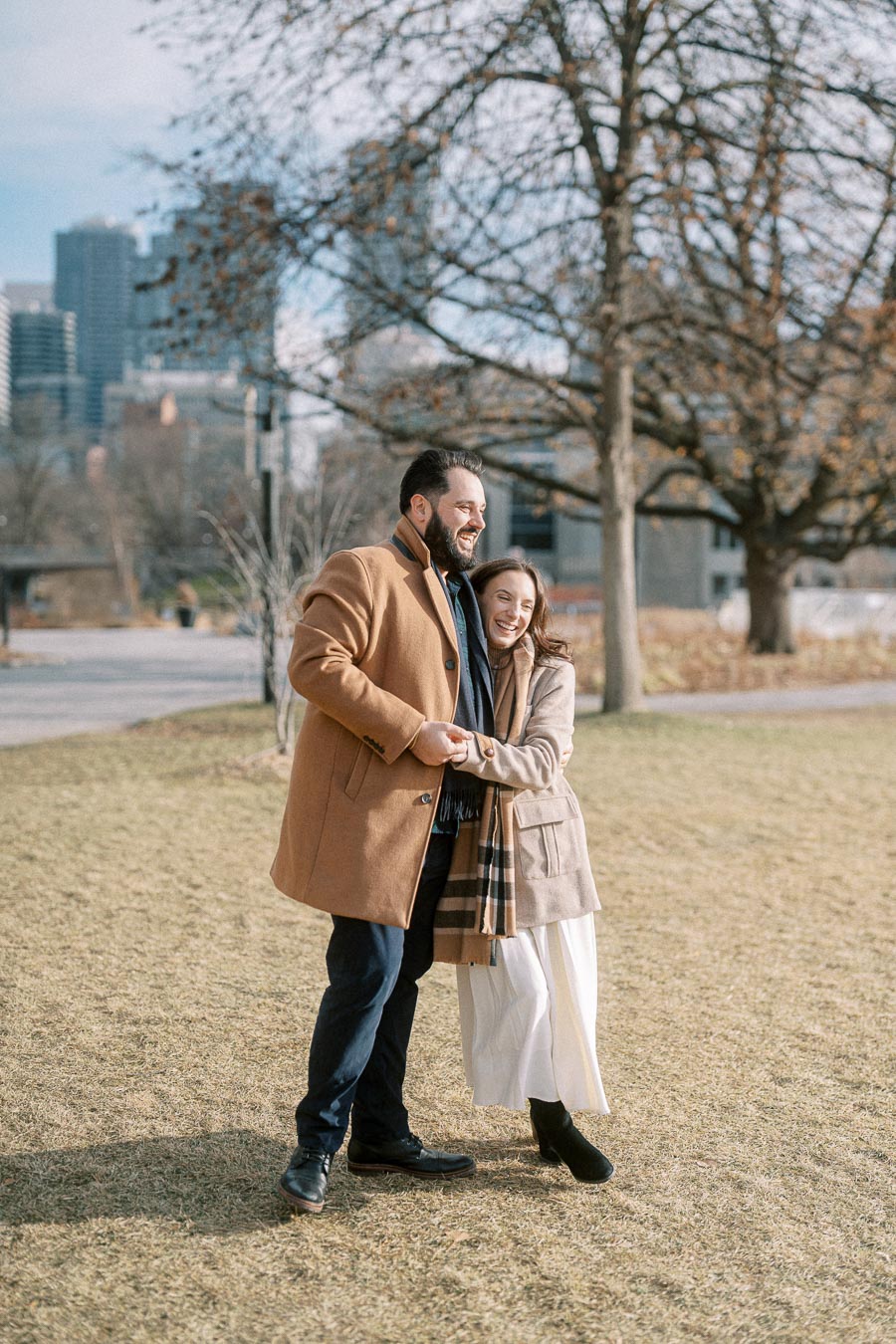 A happy couple enjoying a sunny day in a park, wearing winter coats and scarves, with tall buildings and bare trees in the background.