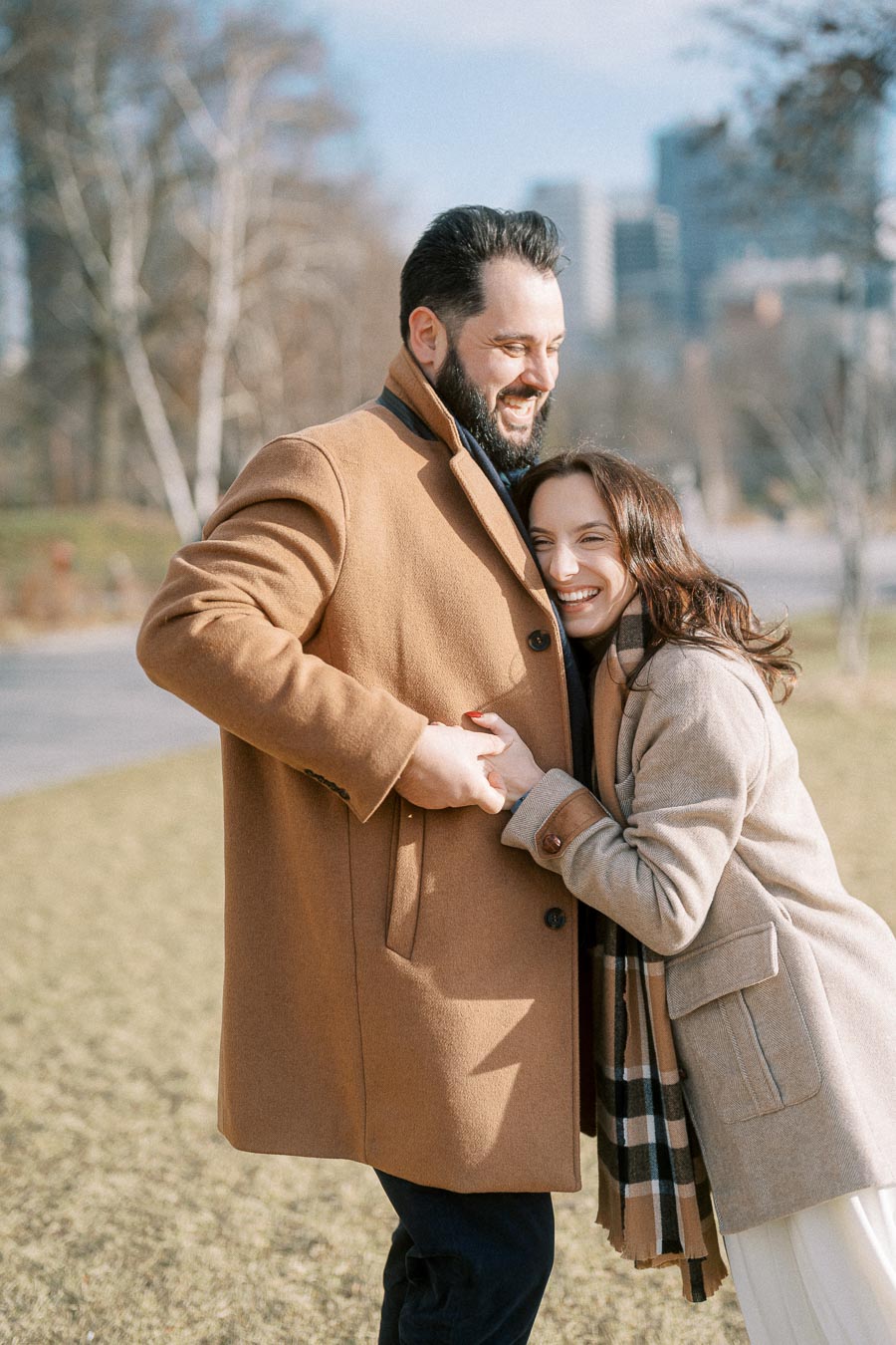 A happy couple embracing in a park on a sunny day, both wearing warm, stylish coats.